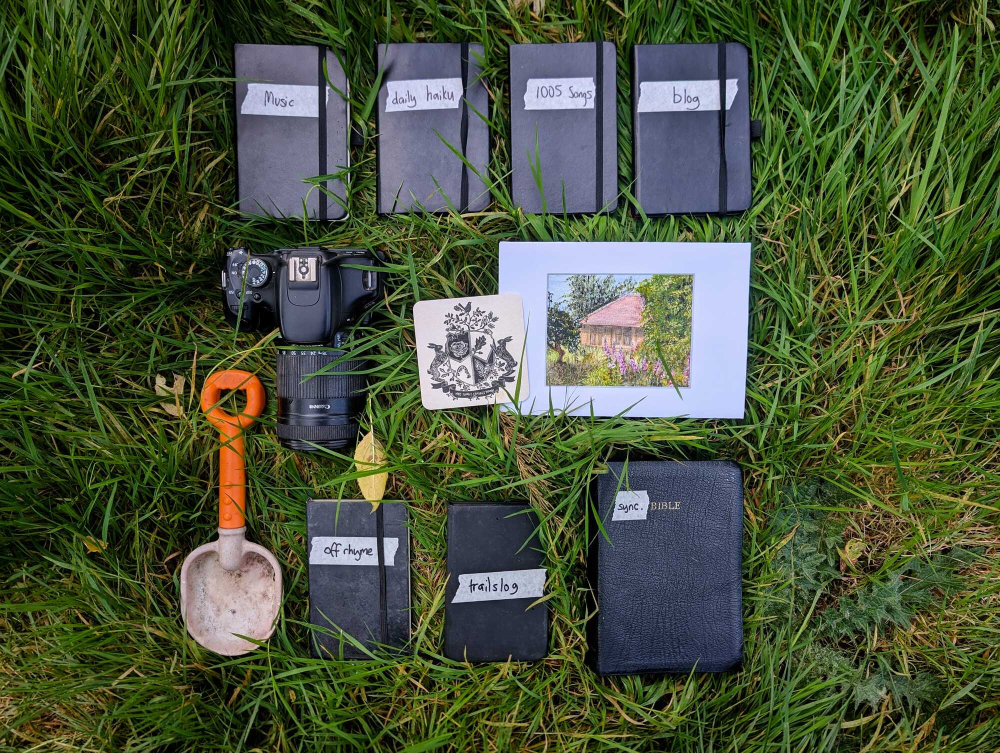Eleven objects laid out on grass: four notebooks labelled in tape with Music, daily haiku, 1005 Songs and blog; a camera; a beer mat printed with a hare crest; a framed painting of a straw-bale building; an orange trowel; notebooks labelled off rhyme and trailslog; and a leather Bible labelled sync.